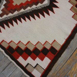 Cream flatweave rug featuring red, brown, and tan geometric borders resting on distressed wood planks.