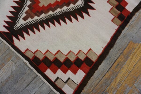 Cream flatweave rug featuring red, brown, and tan geometric borders resting on distressed wood planks.