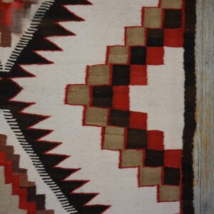A flatwoven rug displaying red, black, tan, and cream geometric patterns on gray weathered wood planks.