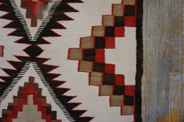 A flatwoven rug displaying red, black, tan, and cream geometric patterns on gray weathered wood planks.