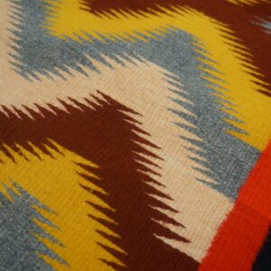 Woven rug featuring thick zigzag stripes in tan, brown, yellow, and gray, with a bright red border detail.