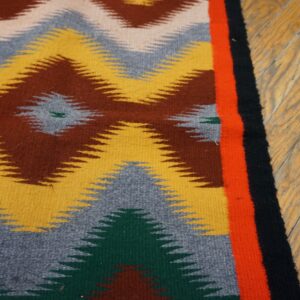 Low-pile rug with geometric zig-zags in yellow, brown, and gray, bordered by orange and black on wood.