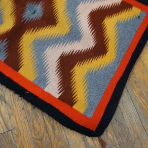 Geometric rug featuring brown, yellow, gray, and green zigzag patterns with red and black borders on wood flooring.