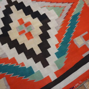 Geometric flat-weave rug with stepped patterns in orange, black, cream, and teal on distressed wood flooring.