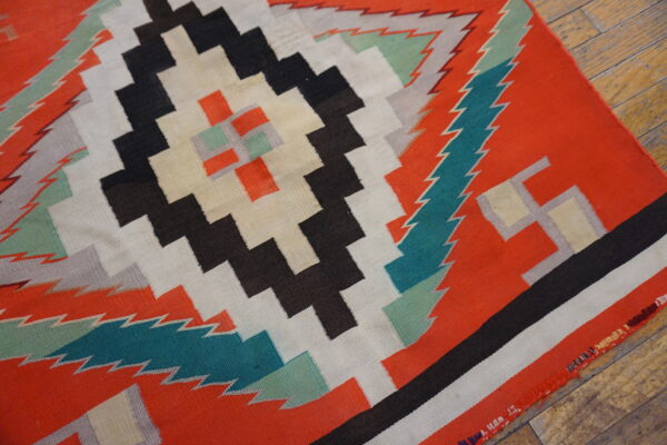 Geometric flat-weave rug with stepped patterns in orange, black, cream, and teal on distressed wood flooring.