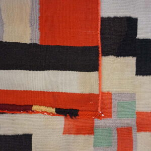 Low-pile flatweave rug detail showing geometric blocks of orange, black, cream, gray, and green.