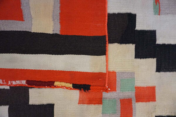 Low-pile flatweave rug detail showing geometric blocks of orange, black, cream, gray, and green.