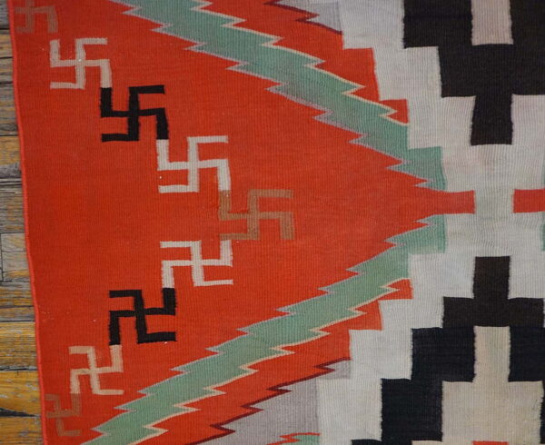 Geometric flatweave rug in orange, black, white, and mint green with repeating hooked cross symbols on wood floors.