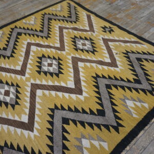 Flatweave rug in gold, black, brown, and grey with a serrated geometric pattern on distressed wood flooring.