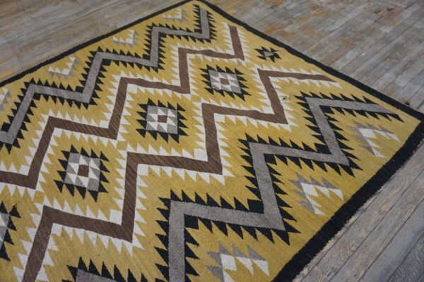 Flatweave rug in gold, black, brown, and grey with a serrated geometric pattern on distressed wood flooring.