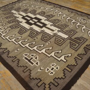 Square flatweave rug featuring geometric gray, olive, cream, and dark brown patterns on distressed wood flooring.