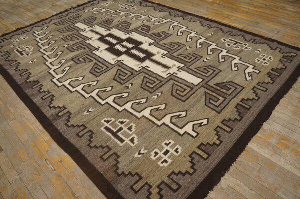 Square flatweave rug featuring geometric gray, olive, cream, and dark brown patterns on distressed wood flooring.