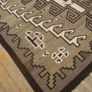 Flatweave rug featuring geometric gray, olive, and off-white patterns rests on distressed wood plank flooring.