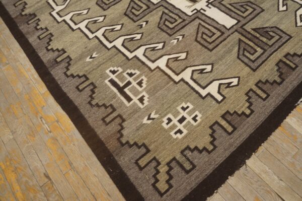 Flatweave rug featuring geometric gray, olive, and off-white patterns rests on distressed wood plank flooring.