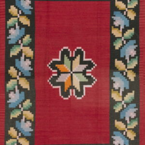 Flat-woven red rug with a dark, pixelated floral border and a multicolored geometric center medallion.