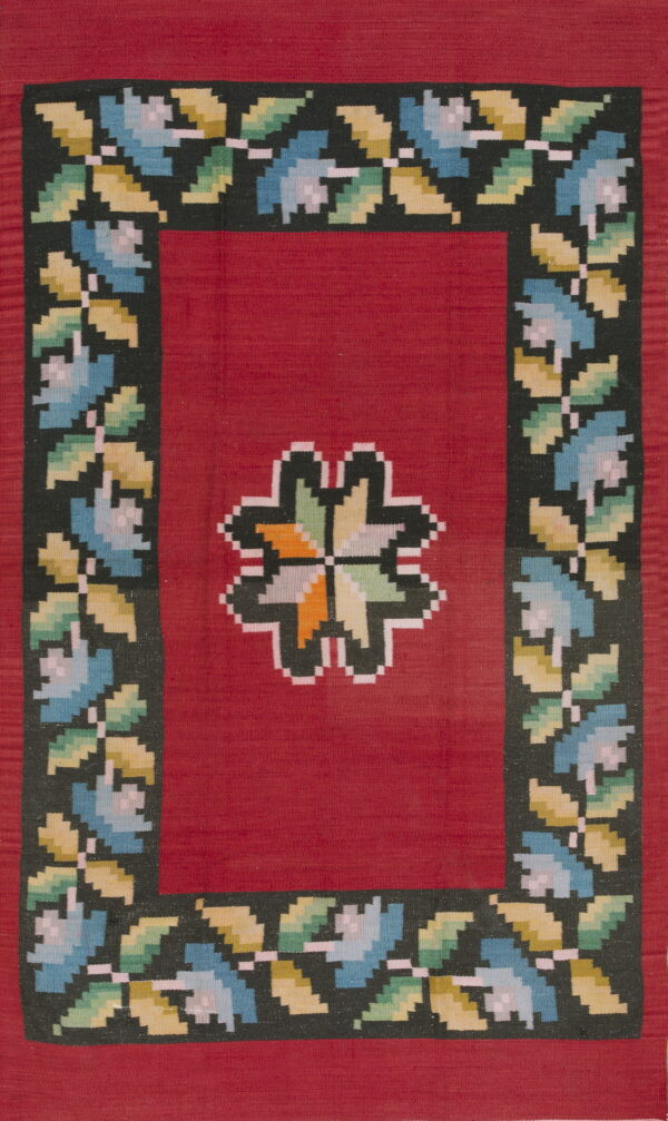 Flat-woven red rug with a dark, pixelated floral border and a multicolored geometric center medallion.