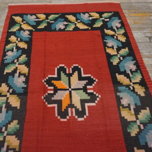 Flatwoven red rug with a central multicolor geometric design and a black floral border on wood floors.