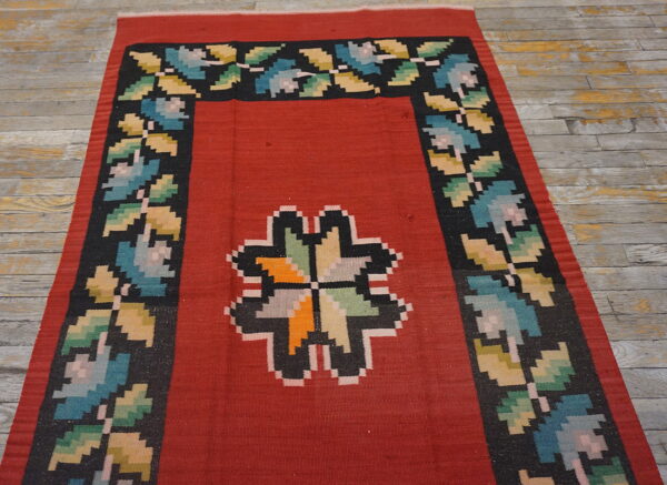 Flatwoven red rug with a central multicolor geometric design and a black floral border on wood floors.