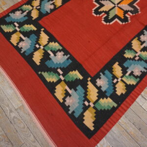 Flatweave rug with a red field and a multicolored, stepped floral border on worn wooden planks.