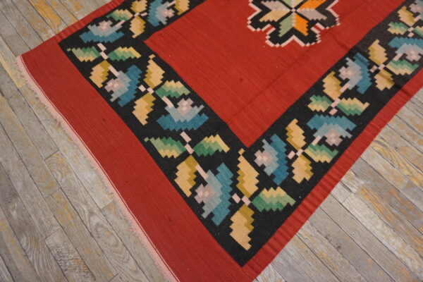 Flatweave rug with a red field and a multicolored, stepped floral border on worn wooden planks.