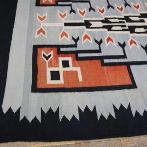 Flatweave rug in black, pale blue, orange, and white geometric patterns rests on distressed wood flooring.