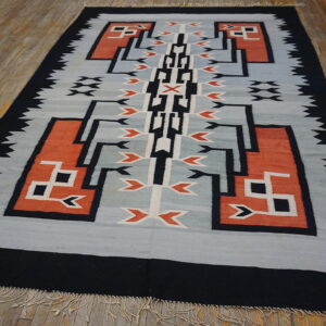 Flatweave rug with a black, orange, white, and gray geometric design on worn hardwood flooring.