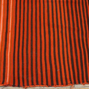 Close-up of a woven rug with thick orange and dark brown vertical stripes on light wooden planks.
