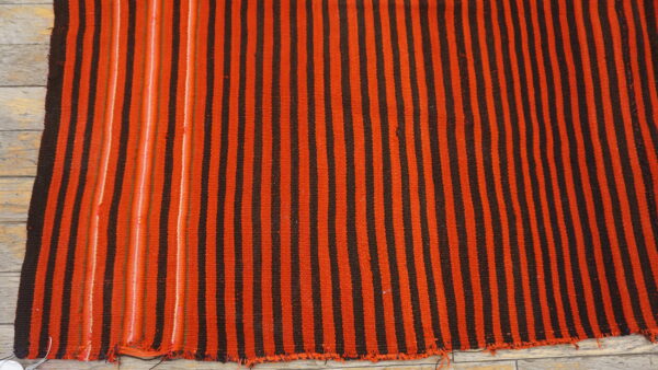 Close-up of a woven rug with thick orange and dark brown vertical stripes on light wooden planks.