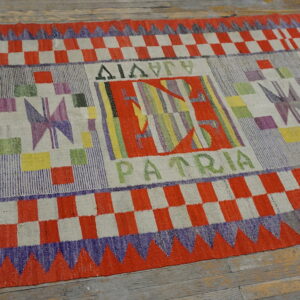 Abstract, multicolored flatweave rug with checkered borders and text resting on worn wooden planks.