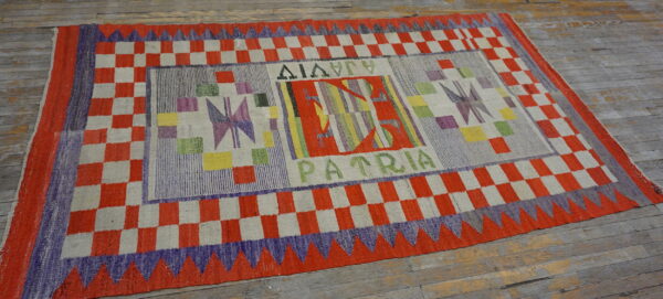 Abstract, multicolored flatweave rug with checkered borders and text resting on worn wooden planks.