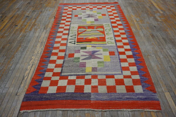 Flatweave runner with red-orange checkerboard borders and multicolored geometric motifs resting on aged hardwood.