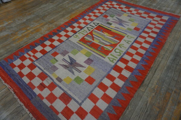 A vibrant flatweave rug with a red checkered border and colorful geometric panels rests on distressed wood flooring.