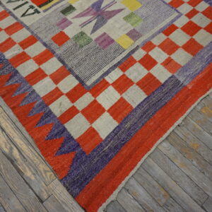 Geometric flatweave rug with a checkerboard border and multiple colors, resting on distressed gray wood flooring.