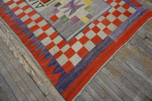Geometric flatweave rug with a checkerboard border and multiple colors, resting on distressed gray wood flooring.