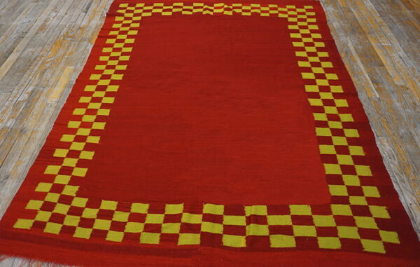 Red flatweave rug with a thick yellow and red checkered border rests on light wood planks.