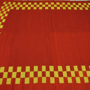 Red flatweave rug featuring a solid central field and a yellow-green checkered border on wood flooring.