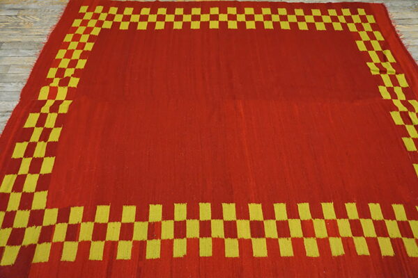 Red flatweave rug featuring a solid central field and a yellow-green checkered border on wood flooring.
