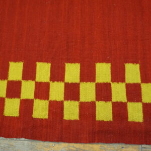 Red and yellow flatweave rug displaying a checkerboard pattern border against rustic wood flooring.