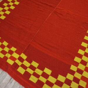 Flat-woven red and yellow rug with a checkered border resting on distressed wood planks.