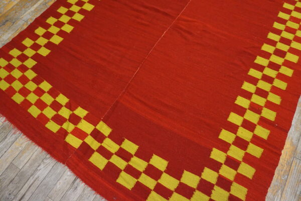 Flat-woven red and yellow rug with a checkered border resting on distressed wood planks.