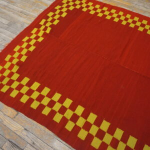 A red and yellow flatweave rug with a checkerboard border lies on worn wooden floorboards.