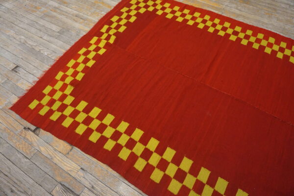 A red and yellow flatweave rug with a checkerboard border lies on worn wooden floorboards.