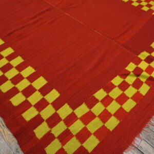 A red flatweave rug with a yellow and red stepped border lies on gray wooden floorboards.