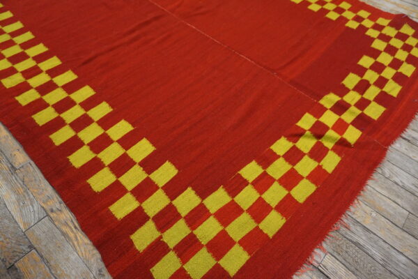 A red flatweave rug with a yellow and red stepped border lies on gray wooden floorboards.