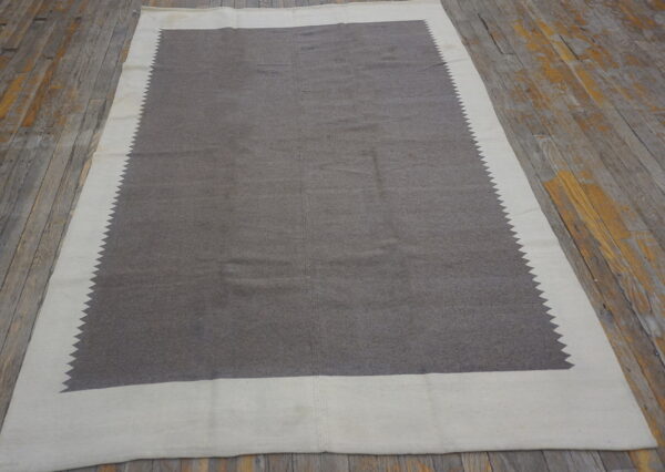 Low-pile gray and off-white rug with a sawtooth inner border rests on distressed wooden floorboards.