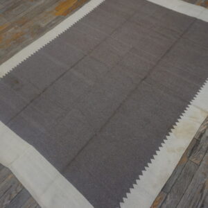 Dark gray-brown flatweave rug with a wide cream border and serrated inner edge on rustic wood floors.