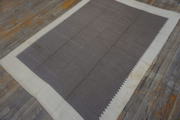 Dark gray-brown flatweave rug with a wide cream border and serrated inner edge on rustic wood floors.