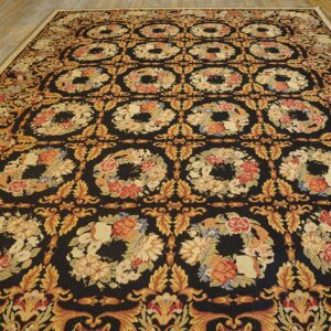 Dark rug with dense repeating floral wreaths in gold, red, and beige on a light hardwood floor.
