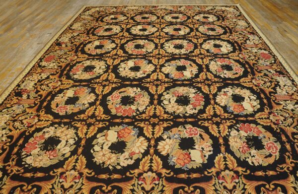 Dark rug with dense repeating floral wreaths in gold, red, and beige on a light hardwood floor.