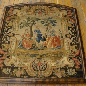 A framed needlepoint tapestry rug featuring a central pastoral scene bordered by dark floral motifs on wood flooring.